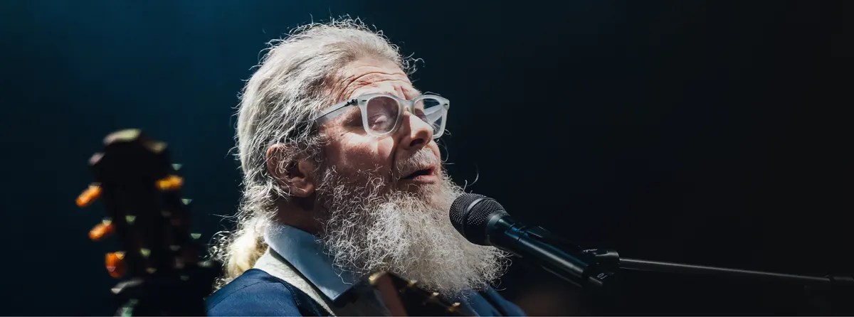 A bearded musician in glasses singing into a microphone, a guitar beside him