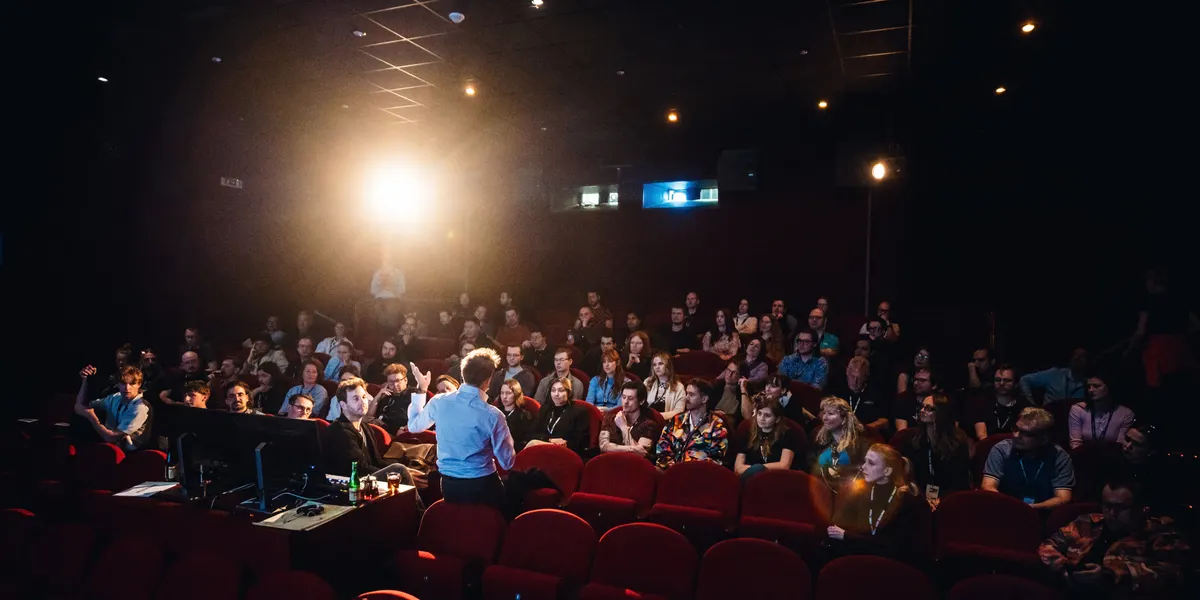 A speaker standing before a full hall at Atlas Cinema, lit from the projection booth