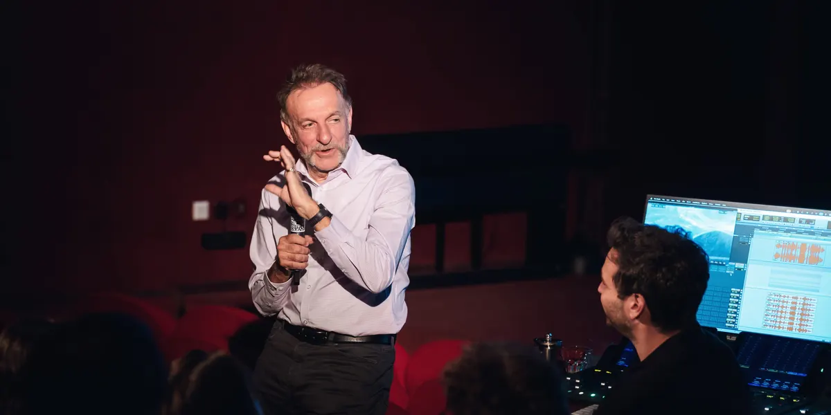 A speaker with a microphone beside a mixing desk and screen in the cinema hall
